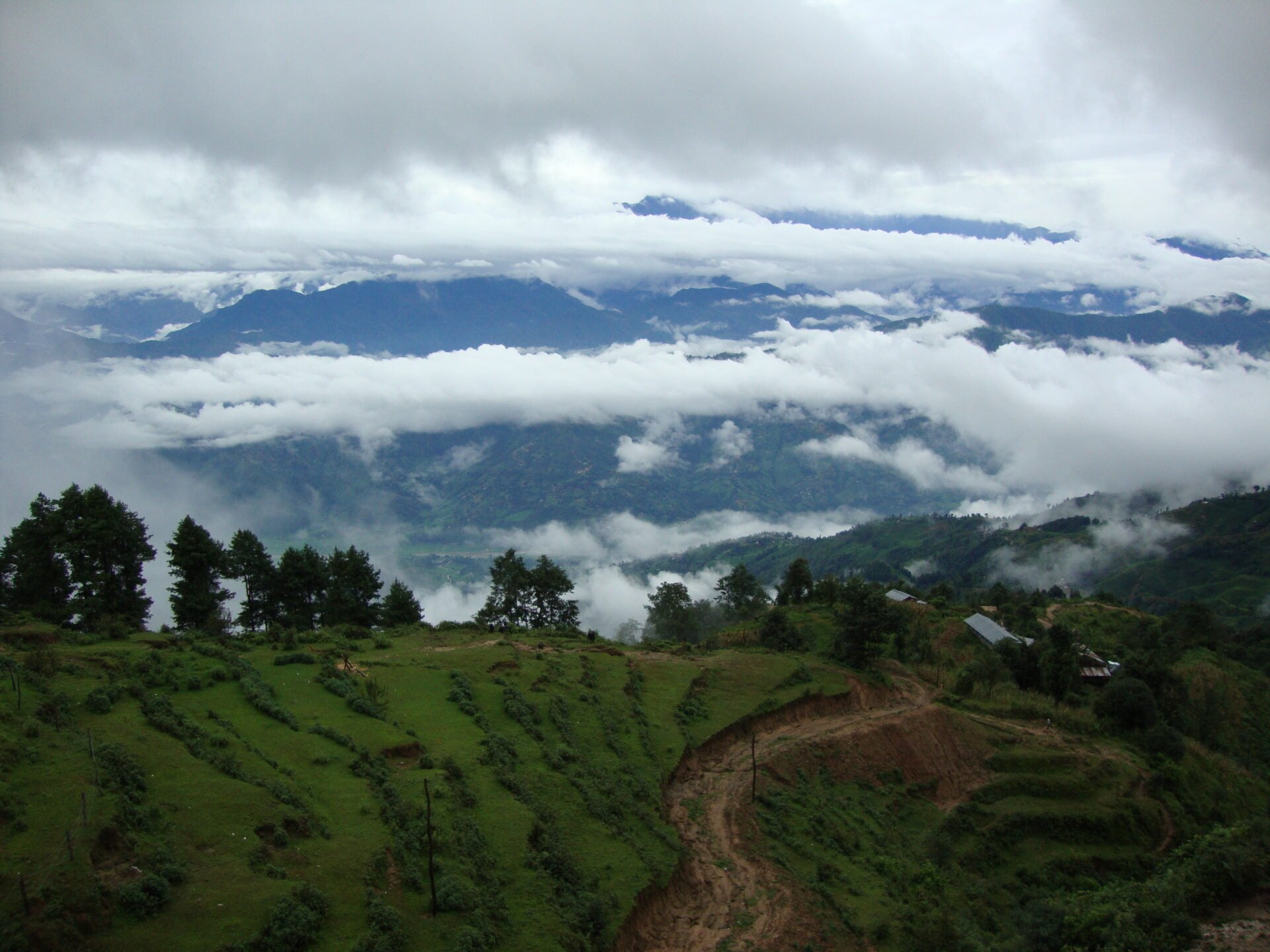 Lush green terraced fields in the foreground with trees scattered throughout, overlooking a valley partially obscured by low-lying clouds, with layers of mountains receding into the background under a cloudy sky.