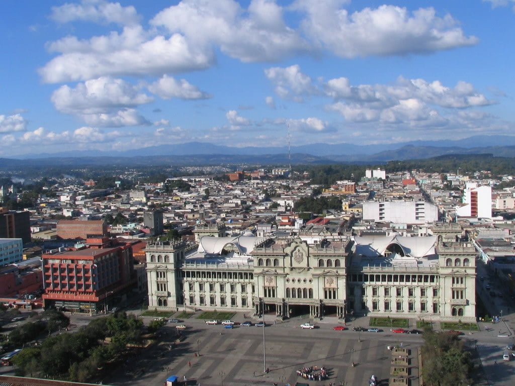 Aerial view of a cityscape with a prominent historic building in the foreground, surrounded by a blend of mid-rise urban structures, expansive blue sky with fluffy clouds above, and distant mountains on the horizon.