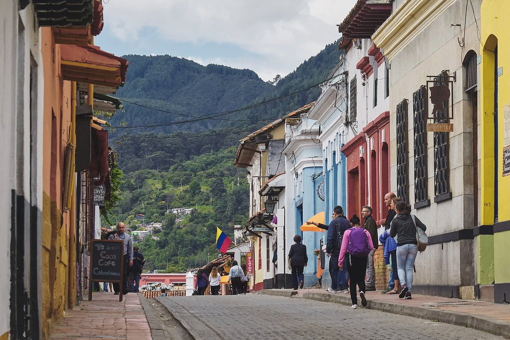 People walking down a cobblestone street in a colorful colonial town with lush green mountains in the background.