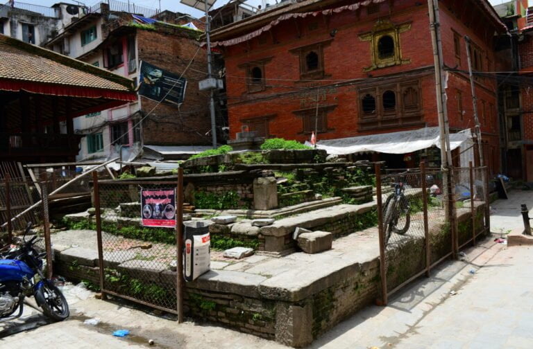 A traditional courtyard in an urban area with historic brick buildings, stone steps, patches of greenery, and a motorcycle parked beside a metal fence.