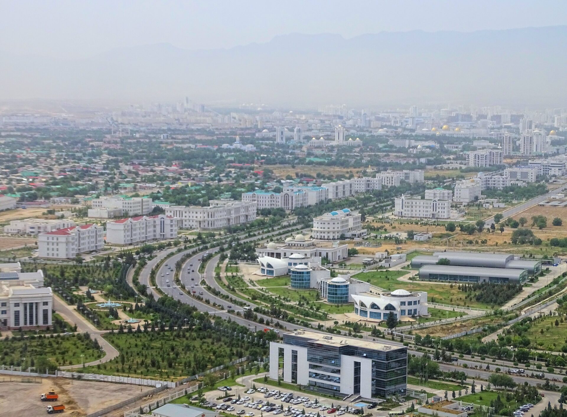 Aerial view of a modern cityscape with a mix of residential and commercial buildings, wide streets with landscaped medians, and distant mountains partially obscured by haze.