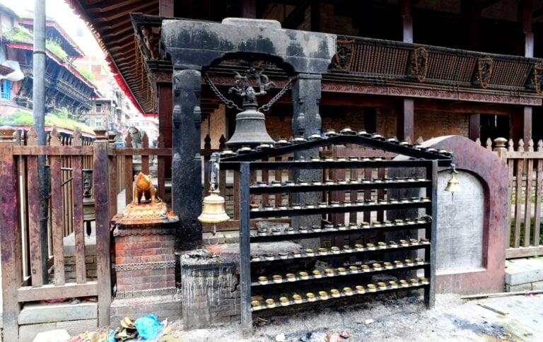 Alt text: A traditional Nepali temple area with a shrine featuring oil lamps, a large bell suspended from a stone structure, and wooden architectural details. A bit of trash is visible on the ground, attesting to the lived-in nature of the space.