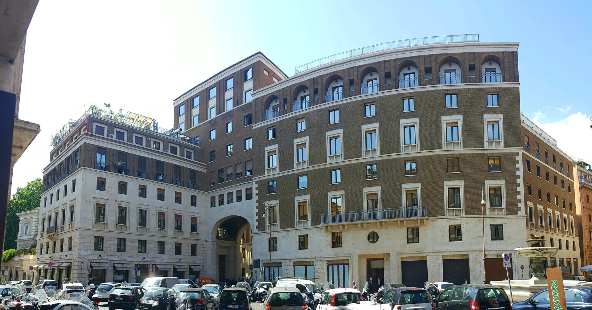Panoramic view of modern urban buildings with various windows, arches, and a visible section of a small fountain, under a clear blue sky. The foreground shows a packed parking area with numerous cars.