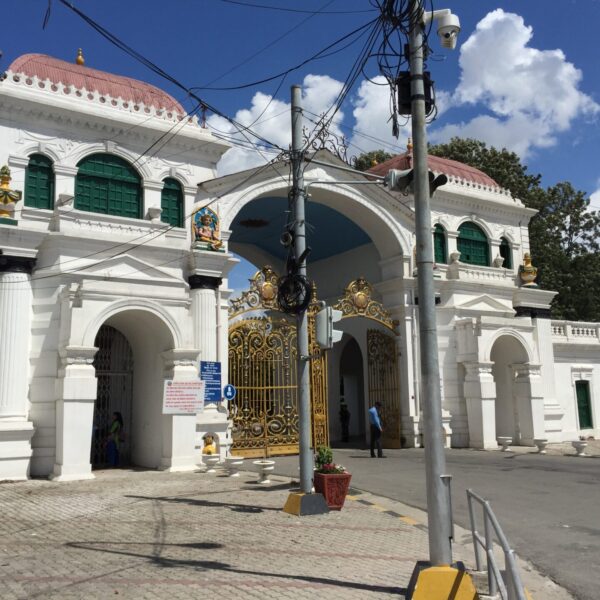An ornate white and gold gate in front of a colonial-style building with dome-shaped roofs and statues on pedestals, under a blue sky with scattered clouds.