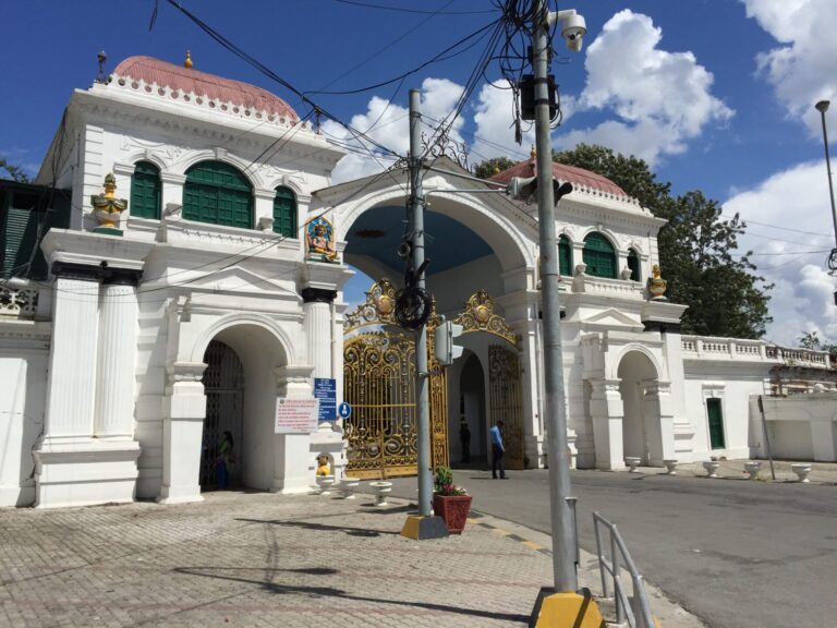 An ornate white and gold gate in front of a colonial-style building with dome-shaped roofs and statues on pedestals, under a blue sky with scattered clouds.