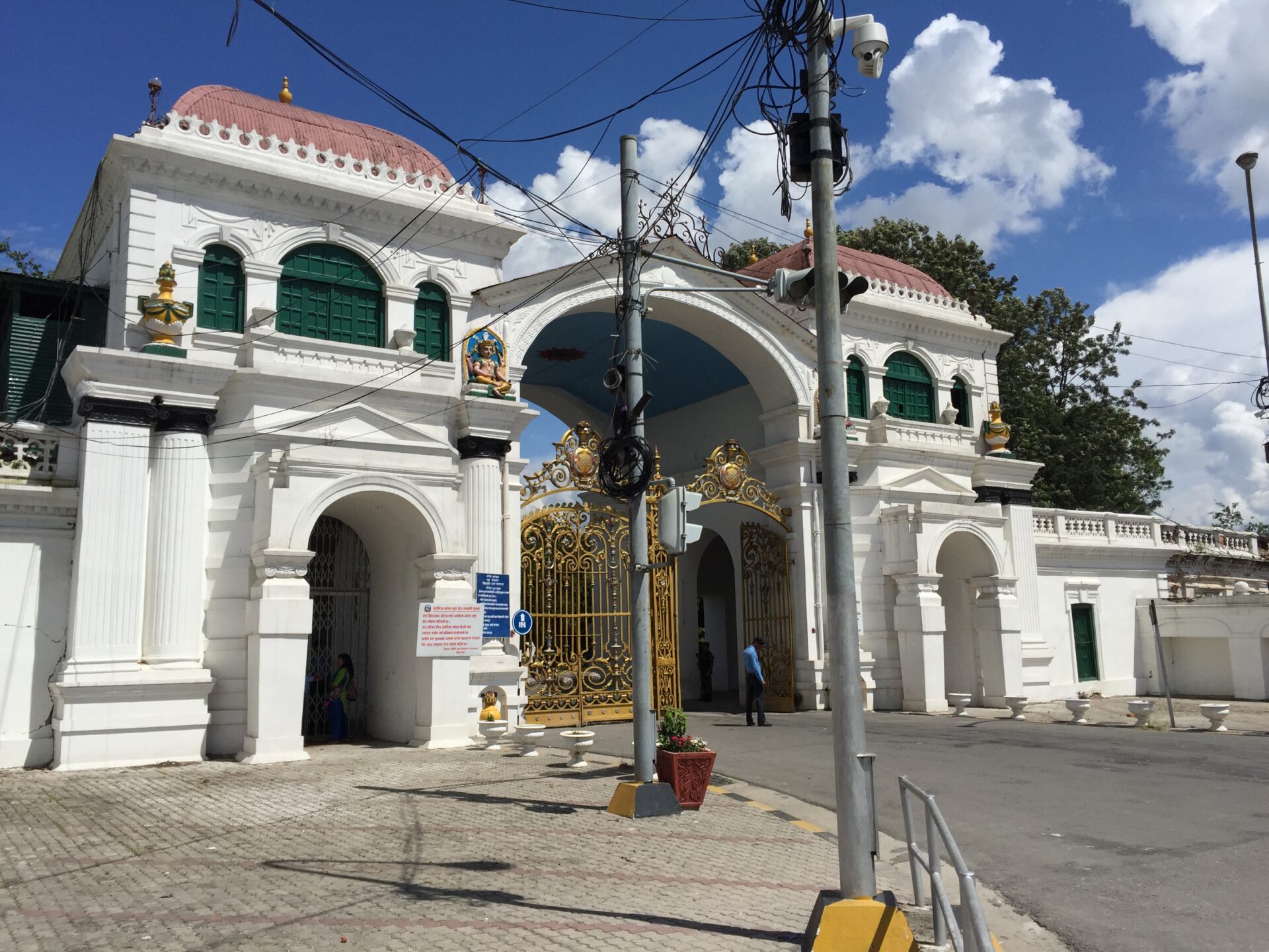 An ornate white and gold gate in front of a colonial-style building with dome-shaped roofs and statues on pedestals, under a blue sky with scattered clouds.