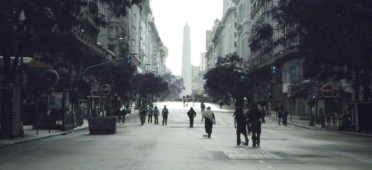 A wide street view with a few pedestrians and a skateboarder, lined with buildings and trees, leading to a distant obelisk monument under overcast skies.