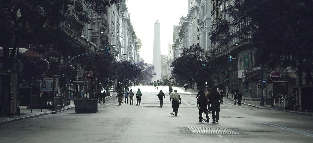 A wide street view with a few pedestrians and a skateboarder, lined with buildings and trees, leading to a distant obelisk monument under overcast skies.