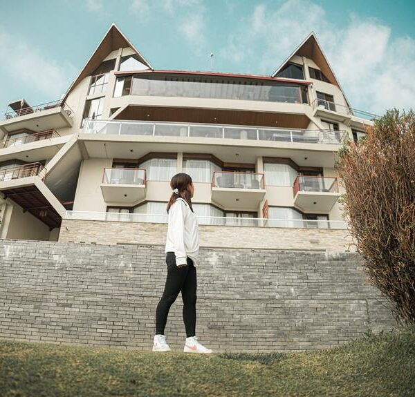 A woman in casual attire stands on a grassy slope in front of a multi-story building with balconies and a clear sky overhead.