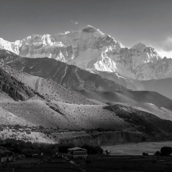 A black and white photo showcasing a layered mountain landscape with a prominent, snow-covered peak in the background, rolling hills in the middle, and a small, solitary building surrounded by flat fields in the foreground.
