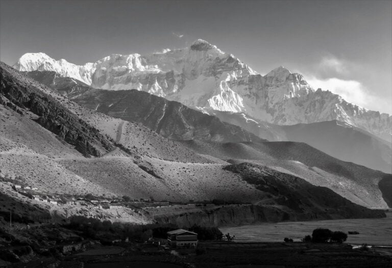 A black and white photo showcasing a layered mountain landscape with a prominent, snow-covered peak in the background, rolling hills in the middle, and a small, solitary building surrounded by flat fields in the foreground.