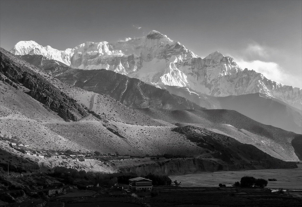 A black and white photo showcasing a layered mountain landscape with a prominent, snow-covered peak in the background, rolling hills in the middle, and a small, solitary building surrounded by flat fields in the foreground.