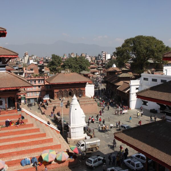 A panoramic view of a bustling square surrounded by traditional Nepalese architecture with tiered temples and pagodas, people mingling around, set against a backdrop of the city with distant mountains under a clear sky.