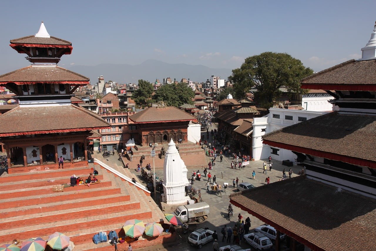 A panoramic view of a bustling square surrounded by traditional Nepalese architecture with tiered temples and pagodas, people mingling around, set against a backdrop of the city with distant mountains under a clear sky.