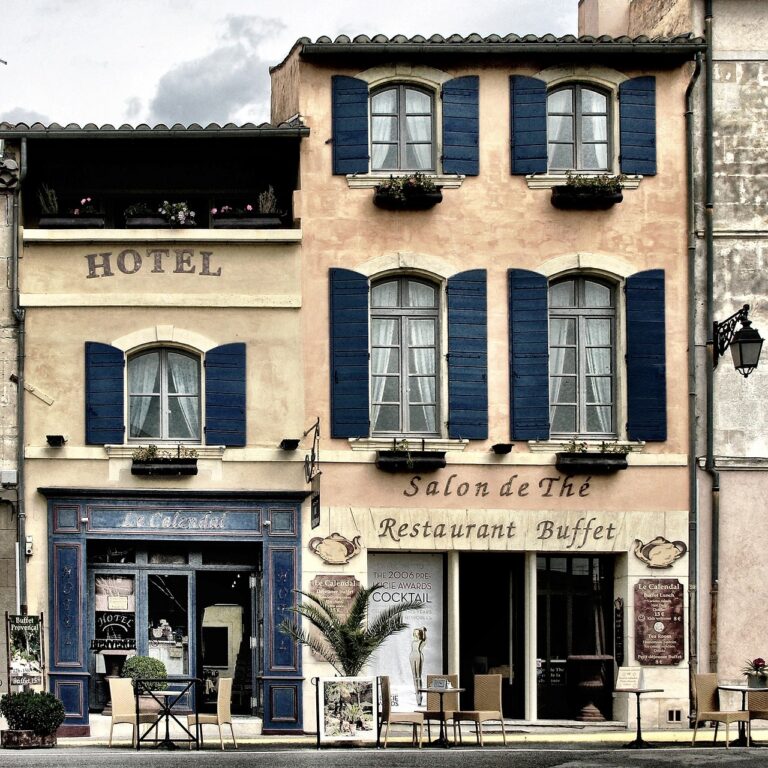Facade of a traditional European hotel with a salon de thé and restaurant below, featuring pastel walls, blue shutters, and a quaint street-side seating area.