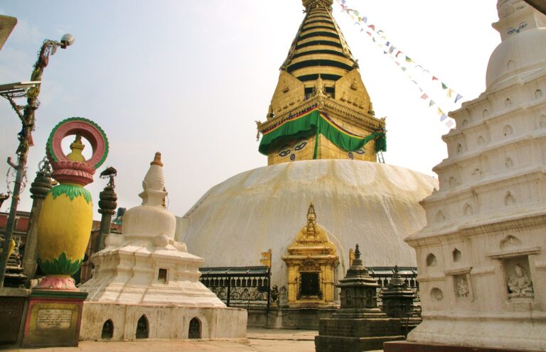 The image shows the Swayambhunath Stupa, also known as the Monkey Temple, in Kathmandu, Nepal, with a large white dome, a golden spire, colorful prayer flags, and smaller surrounding stupas and sculptures.