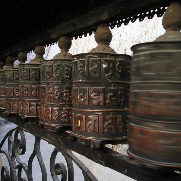 Row of Tibetan prayer wheels mounted on a spindle with carved mantras, one wheel in motion, on a decorative iron railing.