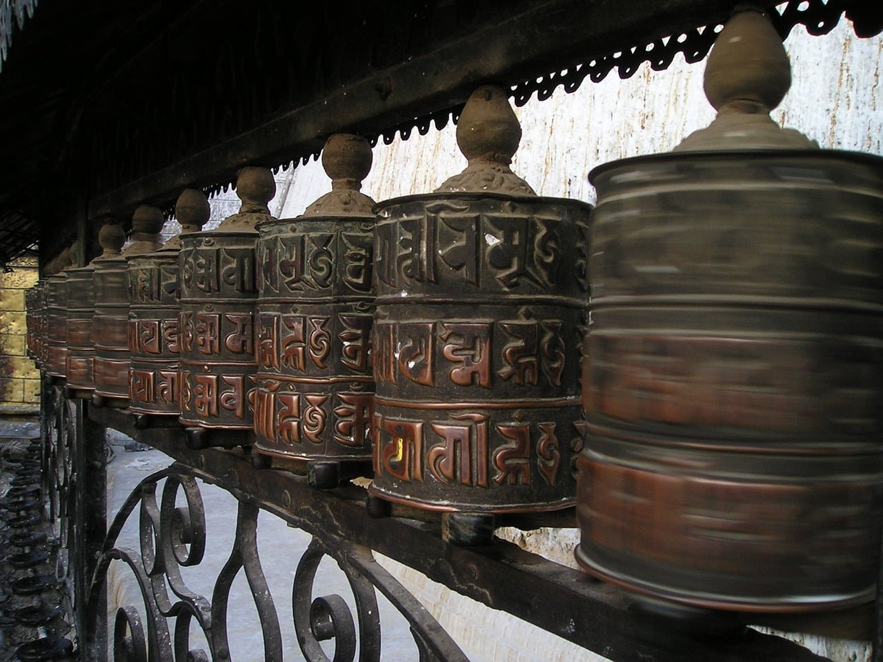 Row of Tibetan prayer wheels mounted on a spindle with carved mantras, one wheel in motion, on a decorative iron railing.