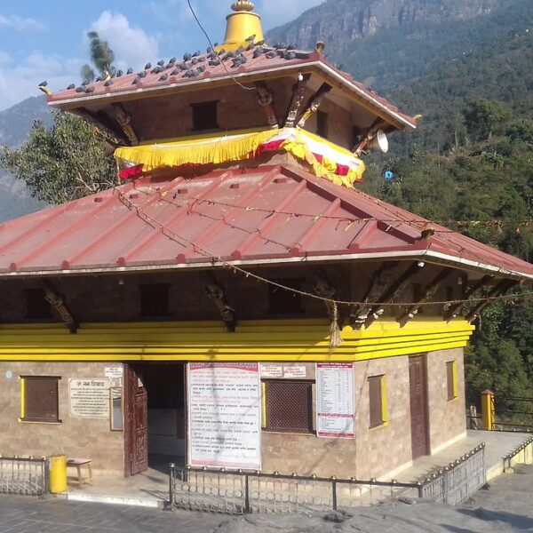 Traditional Hindu temple with a red roof and yellow decorations, perched on a mountainous terrain with a clear blue sky above.