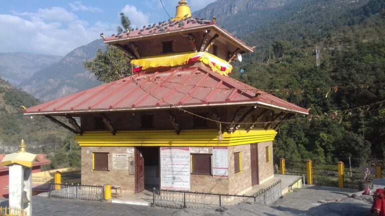 Traditional Hindu temple with a red roof and yellow decorations, perched on a mountainous terrain with a clear blue sky above.