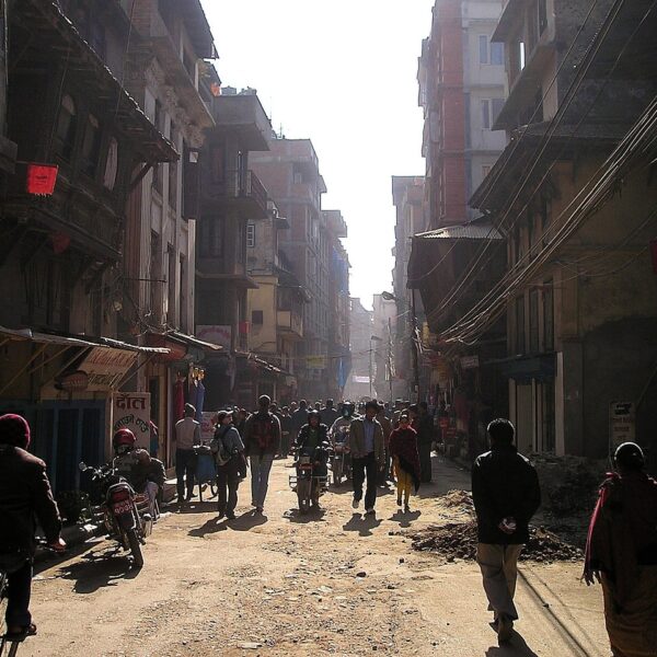 A bustling street scene in a Nepalese urban area filled with pedestrians and motorcyclists, dusty roads, and traditional buildings under hazy sunlight.