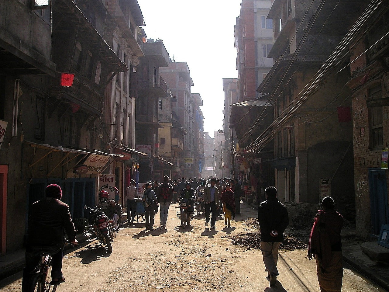 A bustling street scene in a Nepalese urban area filled with pedestrians and motorcyclists, dusty roads, and traditional buildings under hazy sunlight.