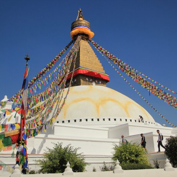 The Boudhanath Stupa under clear blue skies, with colorful prayer flags radiating from the top, and visitors walking around the base.