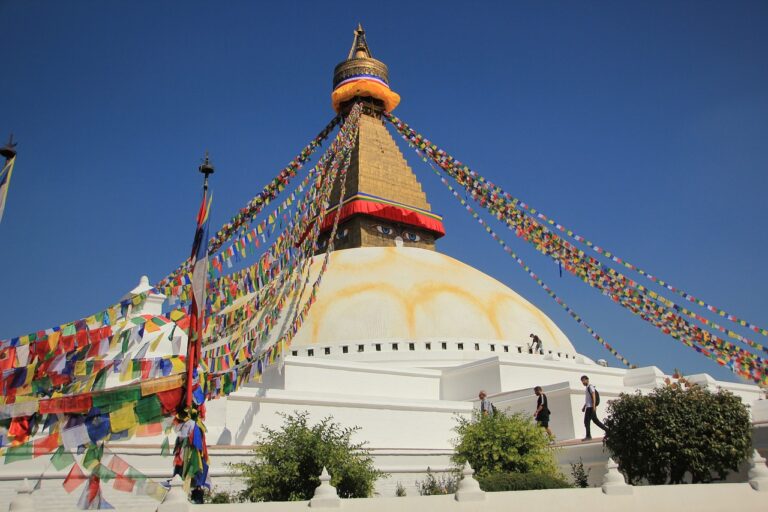 The Boudhanath Stupa under clear blue skies, with colorful prayer flags radiating from the top, and visitors walking around the base.