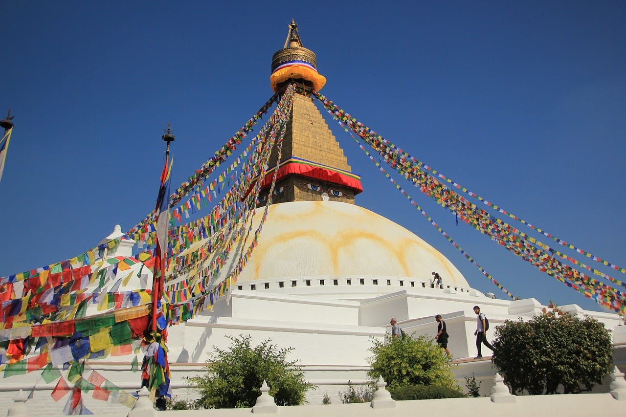 The Boudhanath Stupa under clear blue skies, with colorful prayer flags radiating from the top, and visitors walking around the base.