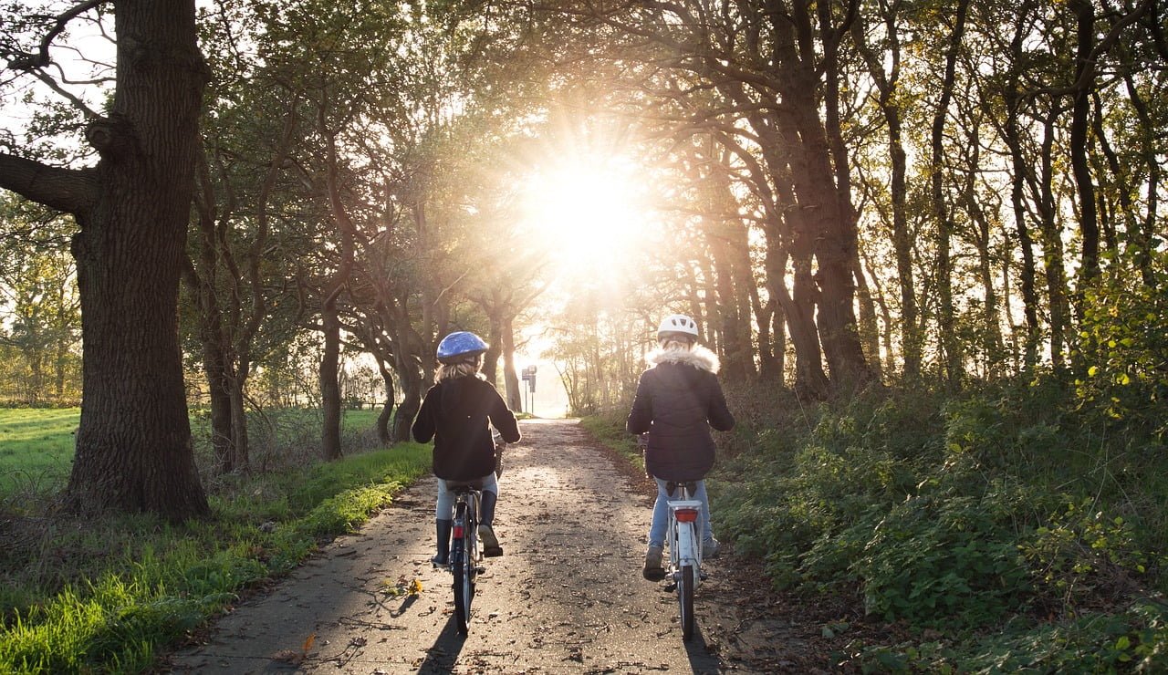 Two individuals wearing helmets cycling on a tree-lined path with the sun setting ahead, creating a bright backlight effect.