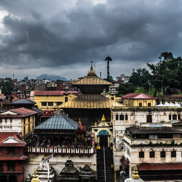 An overcast view of Pashupatinath Temple complex in Kathmandu, Nepal, showcasing its distinctive pagoda-style architecture with visitors gathered on a balcony, backed by a cityscape and surrounding greenery under a cloudy sky.