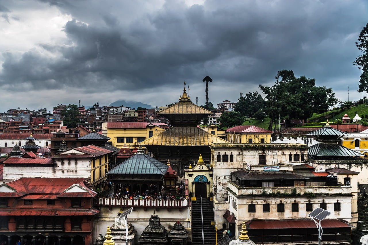 An overcast view of Pashupatinath Temple complex in Kathmandu, Nepal, showcasing its distinctive pagoda-style architecture with visitors gathered on a balcony, backed by a cityscape and surrounding greenery under a cloudy sky.