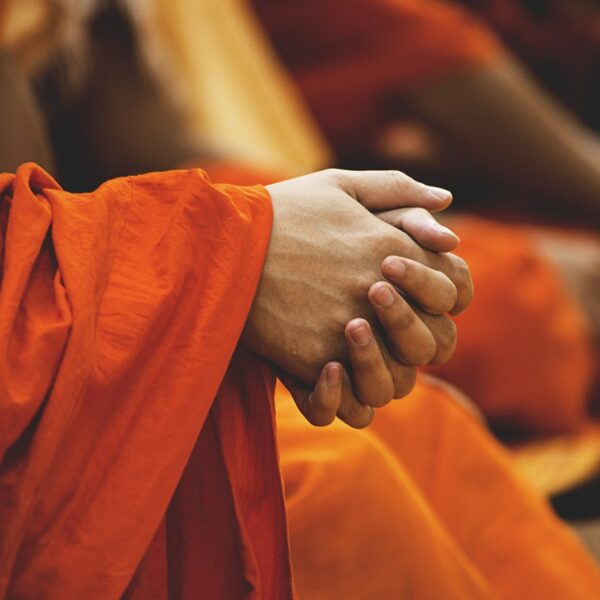 Close-up of clasped hands in focus, draped in vibrant orange fabric, with other similarly dressed individuals blurred in the background.