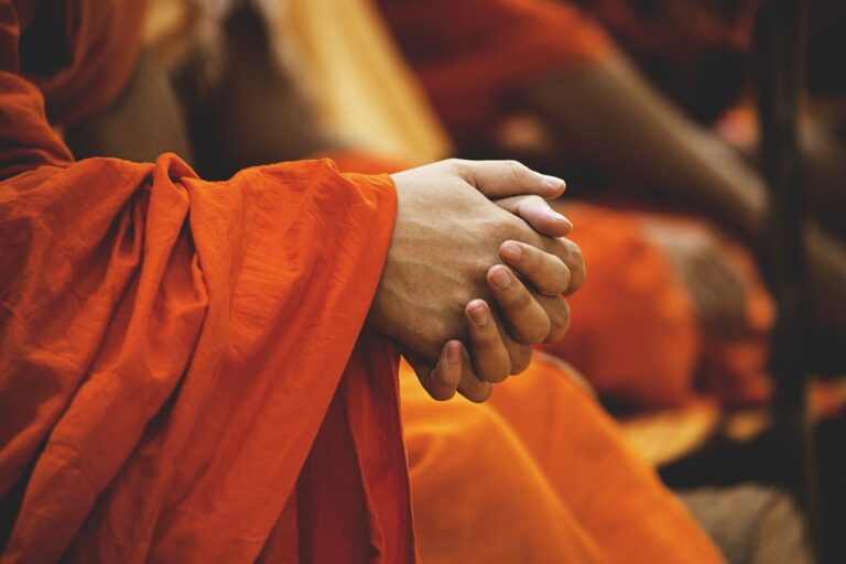 Close-up of clasped hands in focus, draped in vibrant orange fabric, with other similarly dressed individuals blurred in the background.