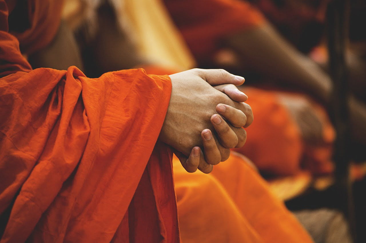Close-up of clasped hands in focus, draped in vibrant orange fabric, with other similarly dressed individuals blurred in the background.