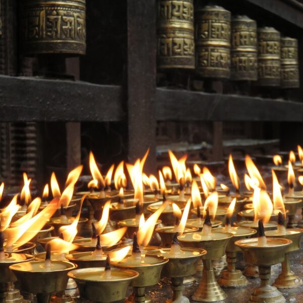 Rows of lit butter lamps with flames in the foreground and Tibetan prayer wheels slightly out of focus in the background.