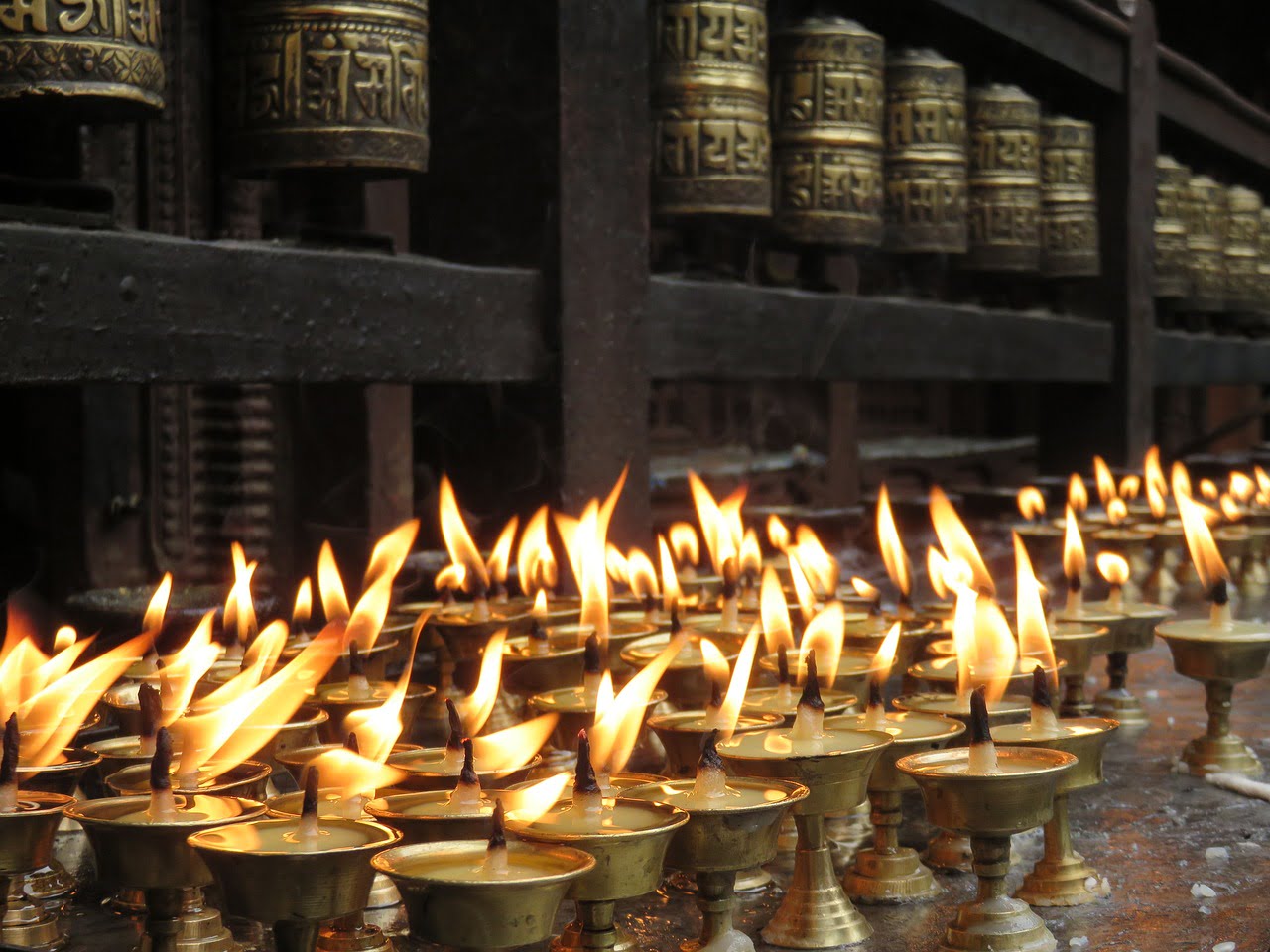 Rows of lit butter lamps with flames in the foreground and Tibetan prayer wheels slightly out of focus in the background.