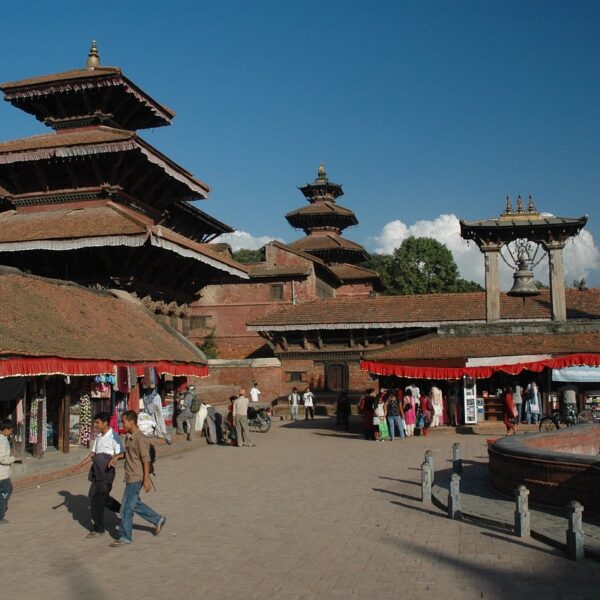 Ancient multi-tiered temples and structures with intricately carved wooden facades at Patan Durbar Square in Nepal, under a clear blue sky, with people walking around and browsing market stalls.