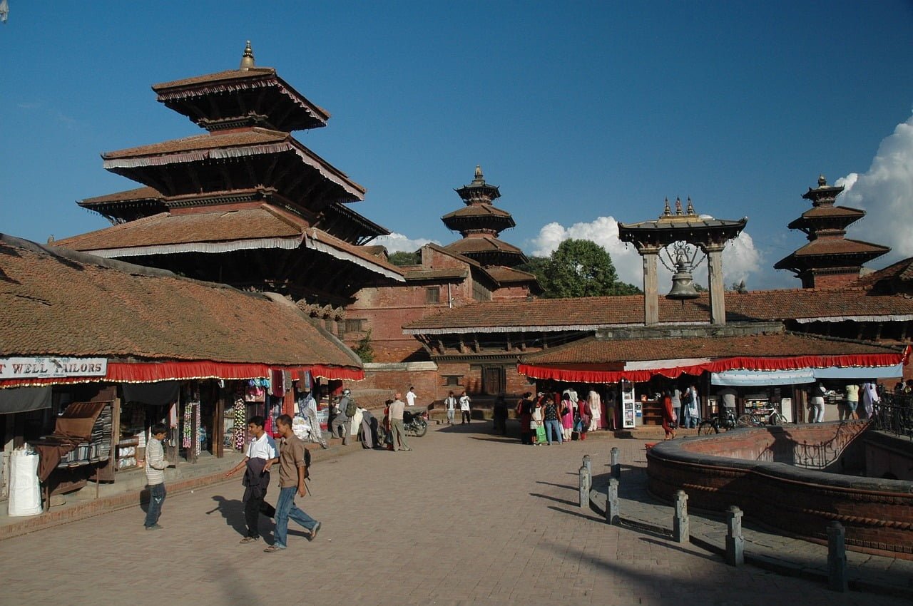 Ancient multi-tiered temples and structures with intricately carved wooden facades at Patan Durbar Square in Nepal, under a clear blue sky, with people walking around and browsing market stalls.