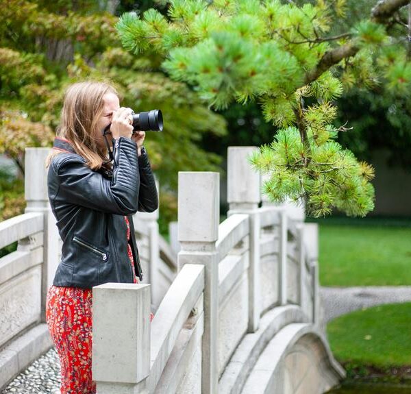 A woman in a black leather jacket and red pants taking a photograph with a DSLR camera while standing on a white bridge in a lush garden setting.