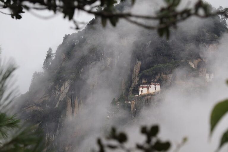 A remote building nestled into the cliffside, partially obscured by mist, with foreground framing by tree branches.