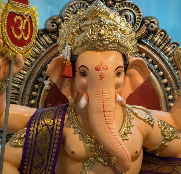 Alt text: A colorful statue of the Hindu deity Ganesha, with an elephant head and four arms, holding a lotus, axe, sweets, and a prayer bead, with the sacred "Om" symbol above the left hand. The deity is ornately adorned with golden headdress and jewelry, set against a blue backdrop.