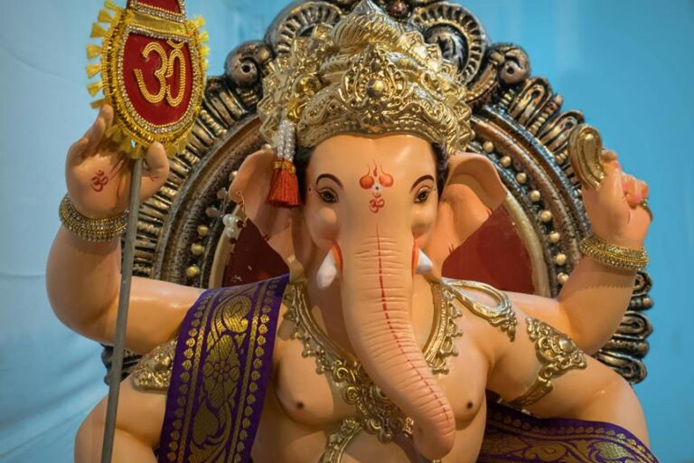 Alt text: A colorful statue of the Hindu deity Ganesha, with an elephant head and four arms, holding a lotus, axe, sweets, and a prayer bead, with the sacred "Om" symbol above the left hand. The deity is ornately adorned with golden headdress and jewelry, set against a blue backdrop.