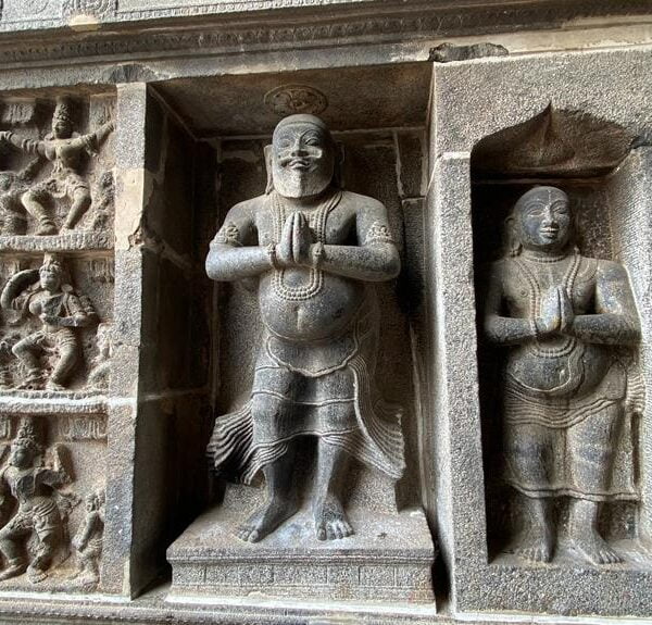 Two ancient stone carvings of human figures in prayer position, set in wall alcoves with intricate relief sculptures on the surrounding stone panels.