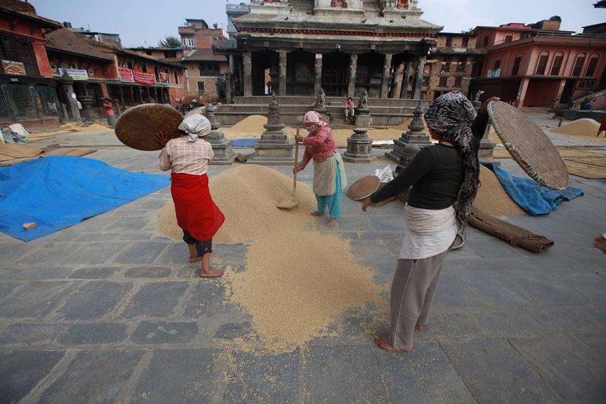 Three women using traditional woven baskets to sift and winnow grain on a stone-paved courtyard in front of an ancient temple with intricate architecture, under a cloudy sky.