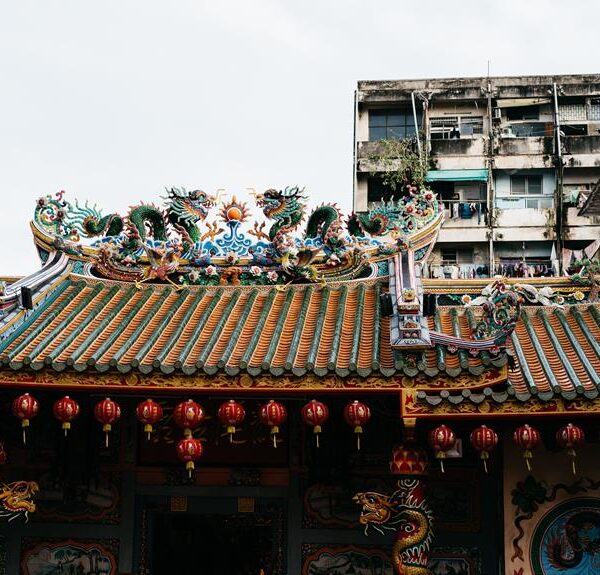 An ornate traditional Chinese temple roof adorned with dragon sculptures and colorful patterns, with red lanterns hanging in front, juxtaposed against a backdrop of an old residential building.