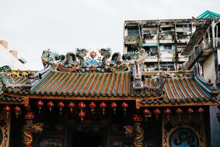 An ornate traditional Chinese temple roof adorned with dragon sculptures and colorful patterns, with red lanterns hanging in front, juxtaposed against a backdrop of an old residential building.