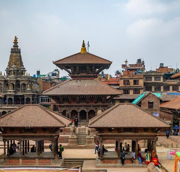 A view of ancient pagoda-style temples and buildings with intricate wood carvings in Patan Durbar Square, Nepal, with people walking and mingling in the square.