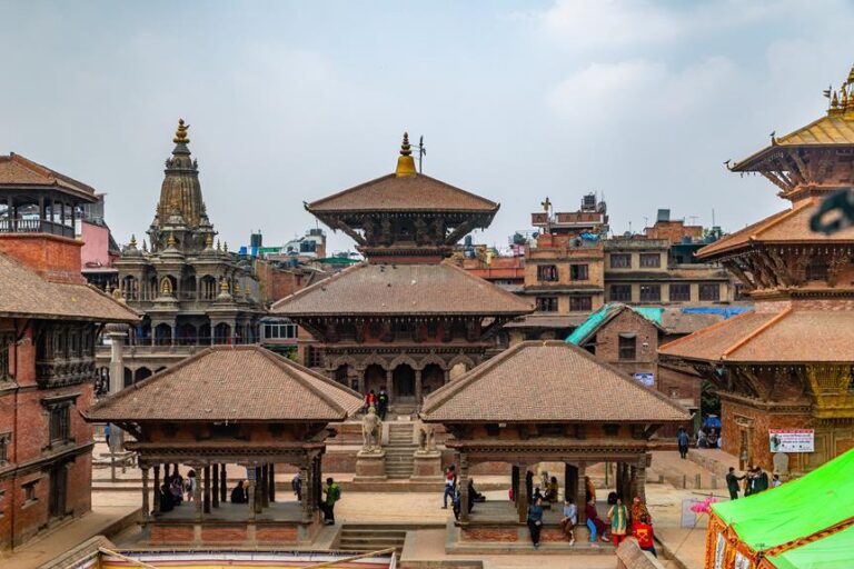 A view of ancient pagoda-style temples and buildings with intricate wood carvings in Patan Durbar Square, Nepal, with people walking and mingling in the square.