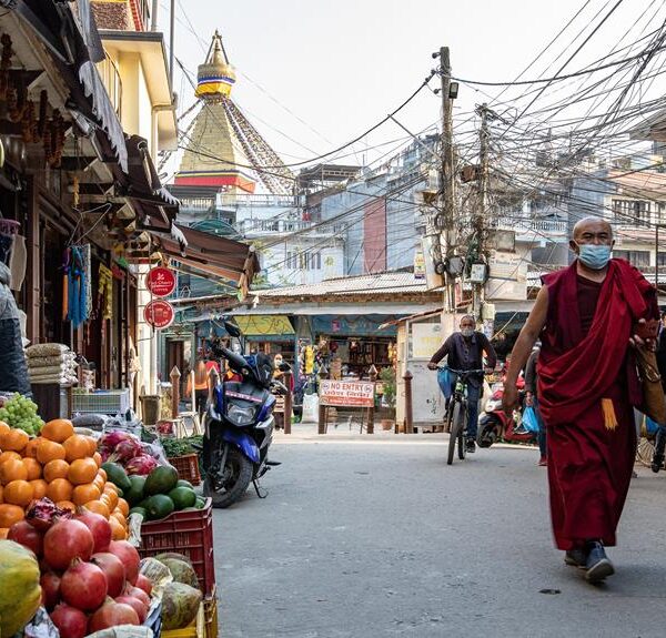 A monk wearing a red robe and a face mask walks down an urban street lined with fruit stalls, a motorcycle, and pedestrians, with a stupa visible in the background and a tangle of electrical wires overhead.